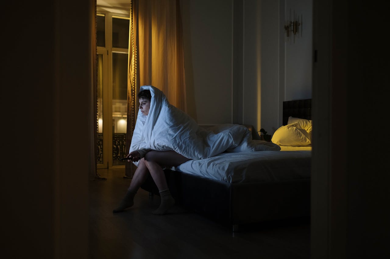 Photo of a Woman Sitting on the Bed while Covered by a White Blanket
